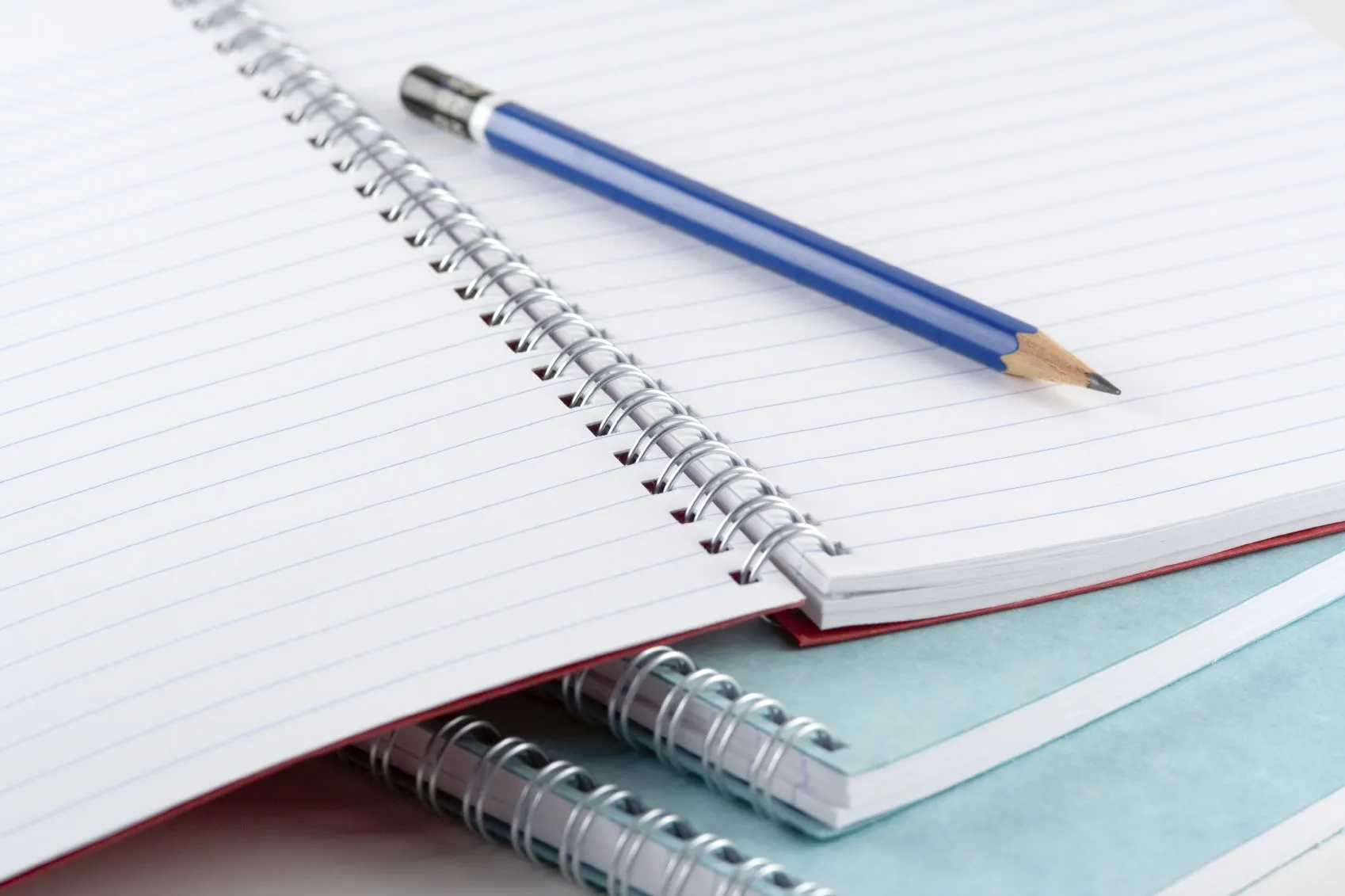 Three spiral bound notebooks laid atop one another, with the one on top open with a pencil laying on it.