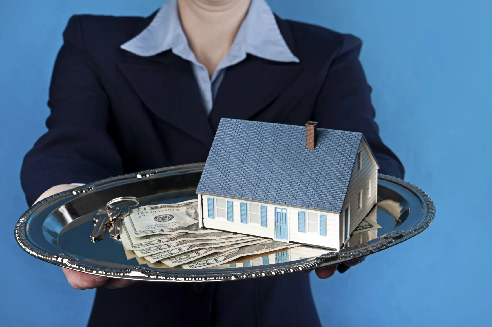 A woman holds an offering plate out with money, a model house, and a set of keys on it.