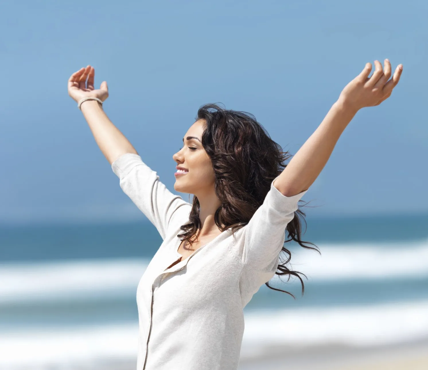 A woman on the beach with her arms outstretched upward, wearing a white blouse.