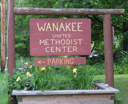 A wooden sign that reads Wanakee United Methodist Center, and a smaller attached sign that reads Parking with an arrow, with a flower box below the signs with trees and grass in the background