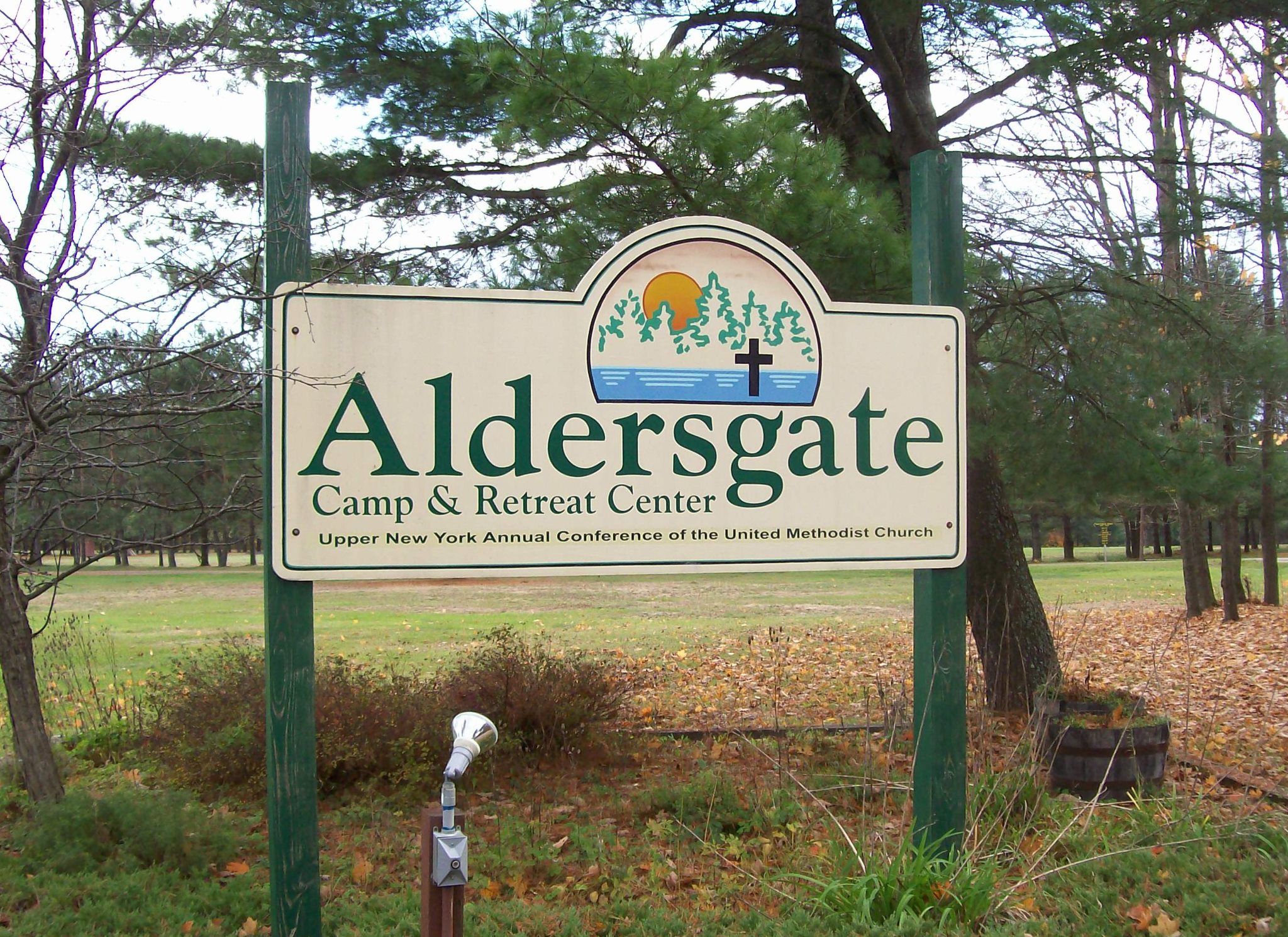 a sign that reads Aldersgate Camp & retreat center, upper new york annual conference of the United Methodist Church, with a backround of an open field and trees in fall