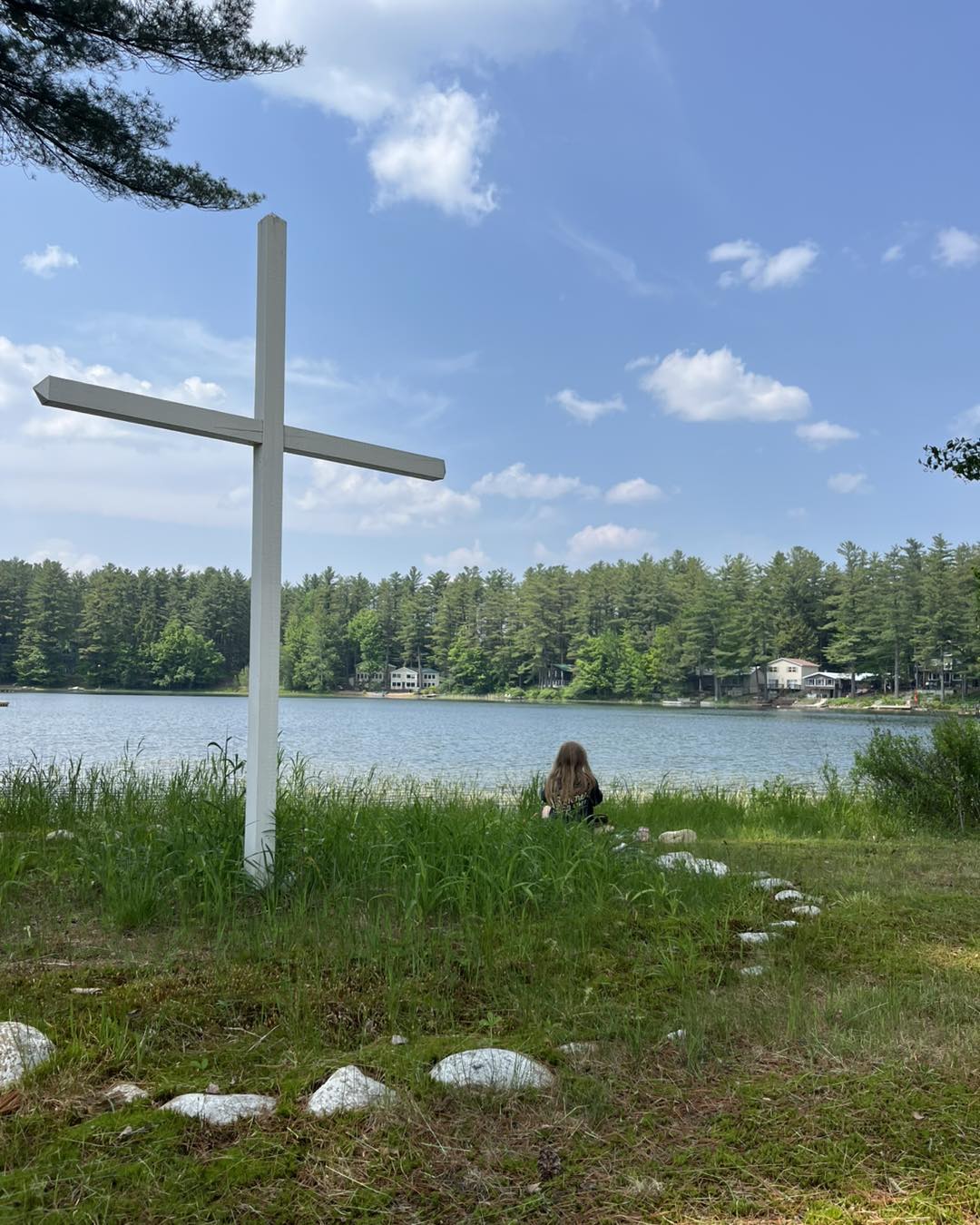 A white cross on the bank of a lake with trees and blue skies in the background