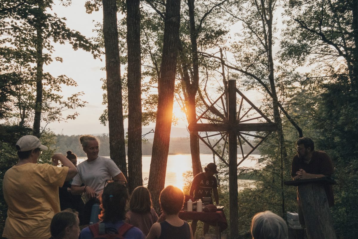 Group of people gathered in a wooded area seated on pews with a large wooden cross at the front looking over a lake at sunset