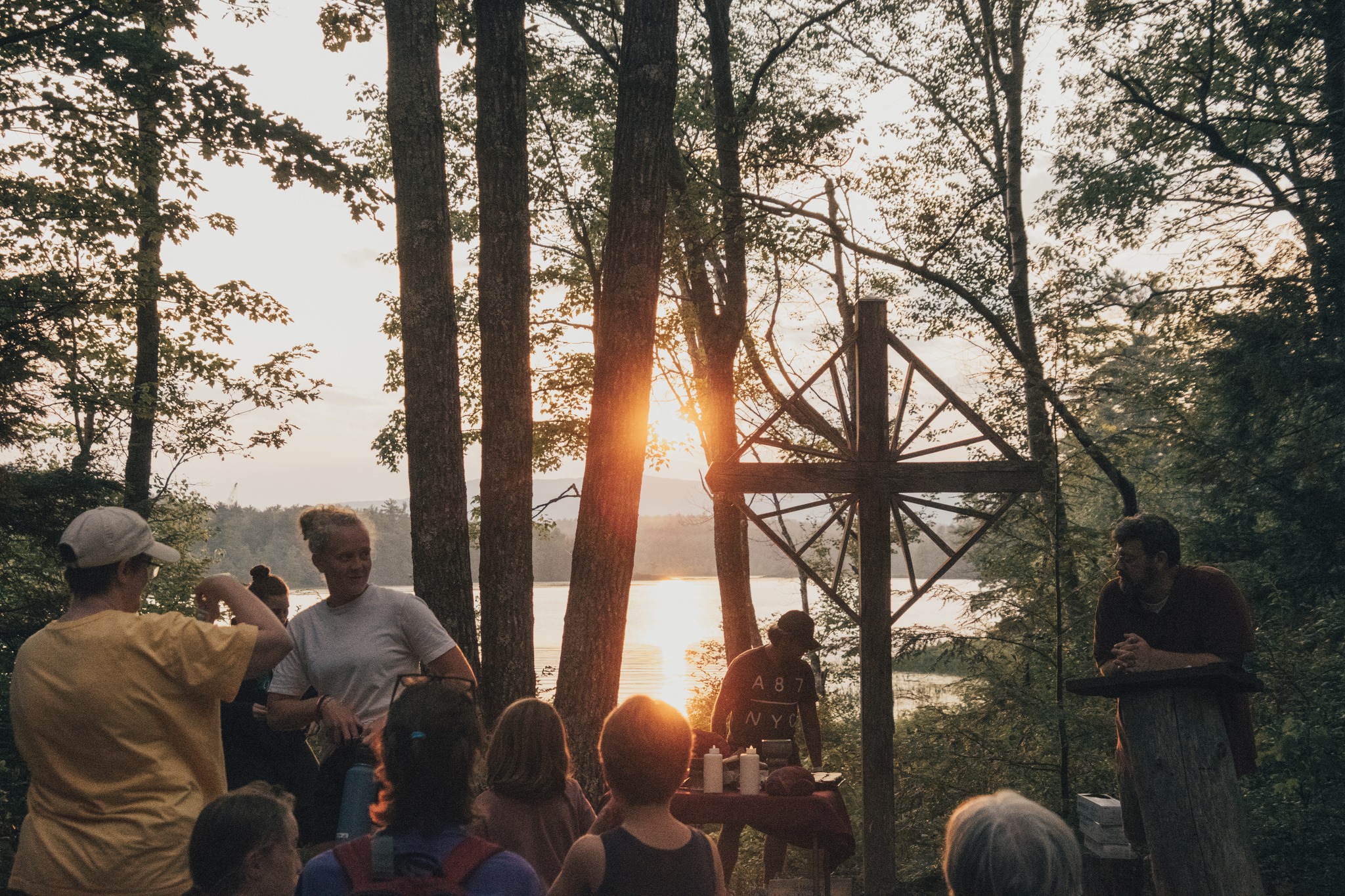 Group of people gathered in a wooded area seated on pews with a large wooden cross at the front looking over a lake at sunset