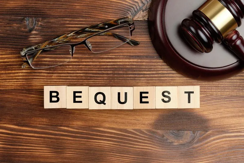 Scrabble tiles on a wood table spelling the word "Bequest", with glasses and a gavel resting above them.