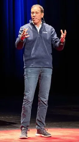 A man giving a speech on a stage in front of a blue curtain.