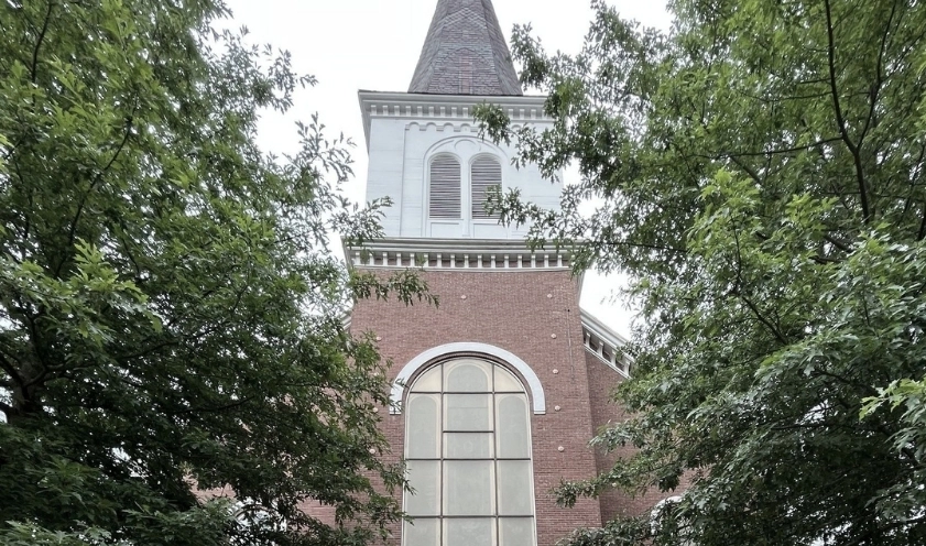 A large brick spire of a church stands between thick, leafy tree canopies