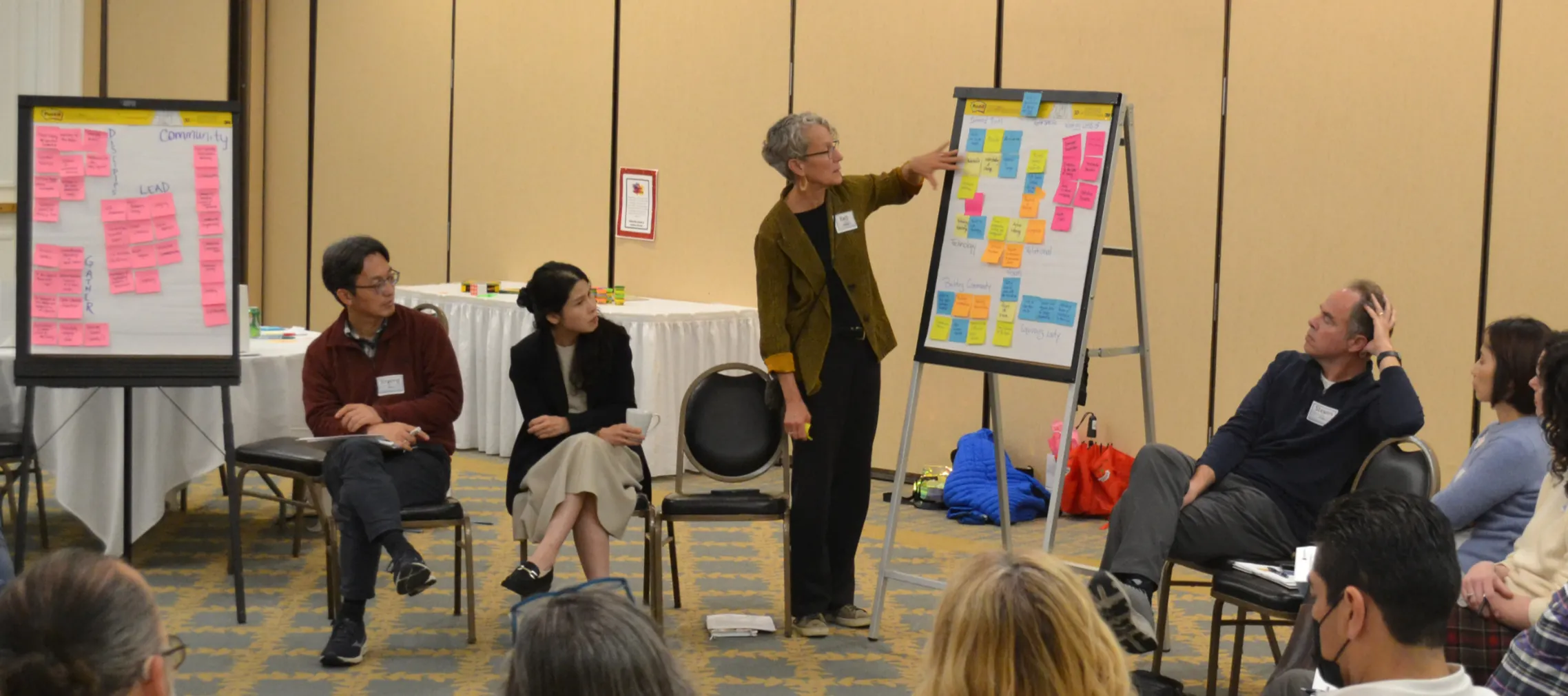 A woman standing in front of an easel with colorful post-it notes speaking to a group of people in a conference room seated in chairs