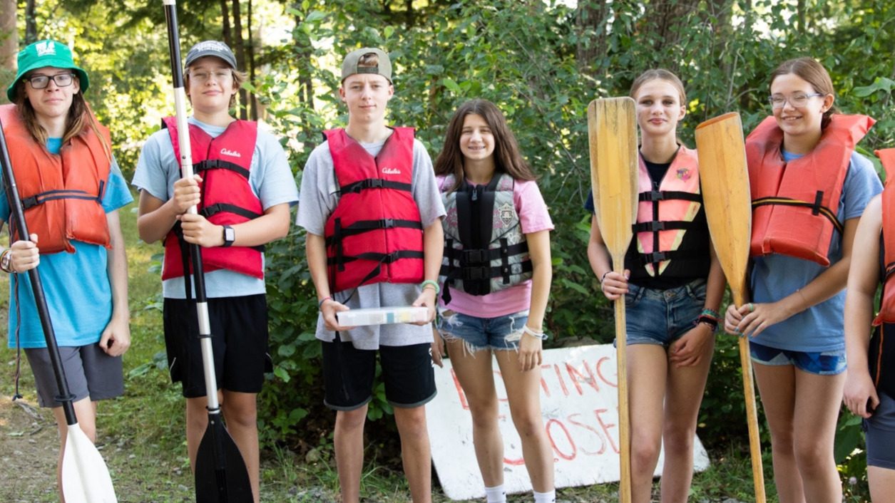 A group of kids in life jackets holding kayak and canoe oars posing for a photo outside