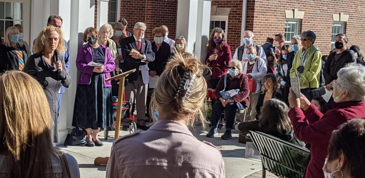 A large crowd of people making a circle around a speaker holding a paper and using a microphone outside of a building