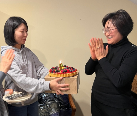 A woman holds a fruit tart with a birthday candle lit and another woman stands with her hands clasped in anticipation to blow out the candle