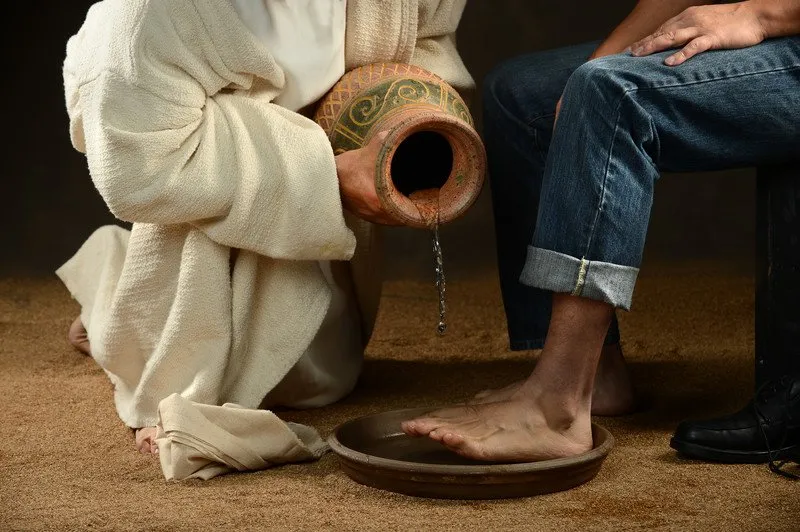 A priest in white robes washes the feet of a person sitting in a chair.