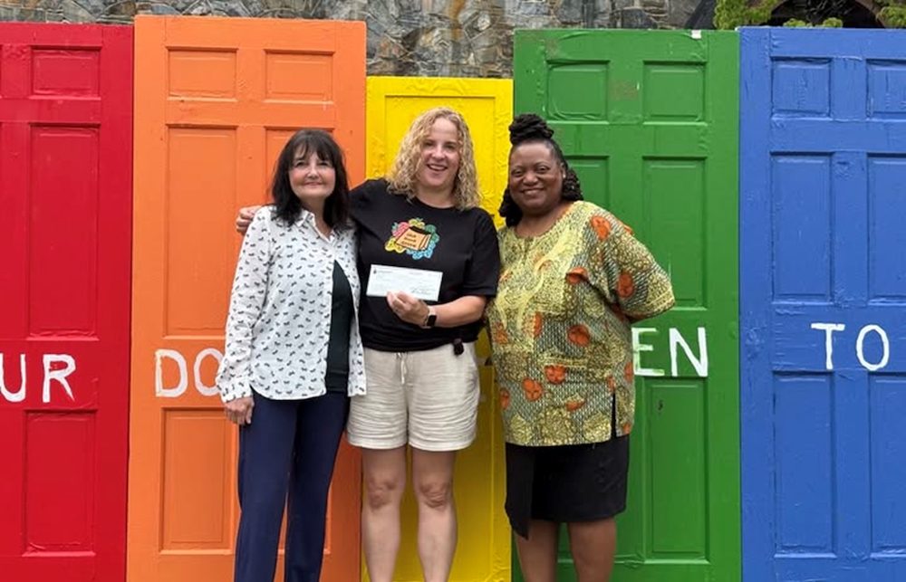 Three women stand in front of rainbow colored doors holding a check