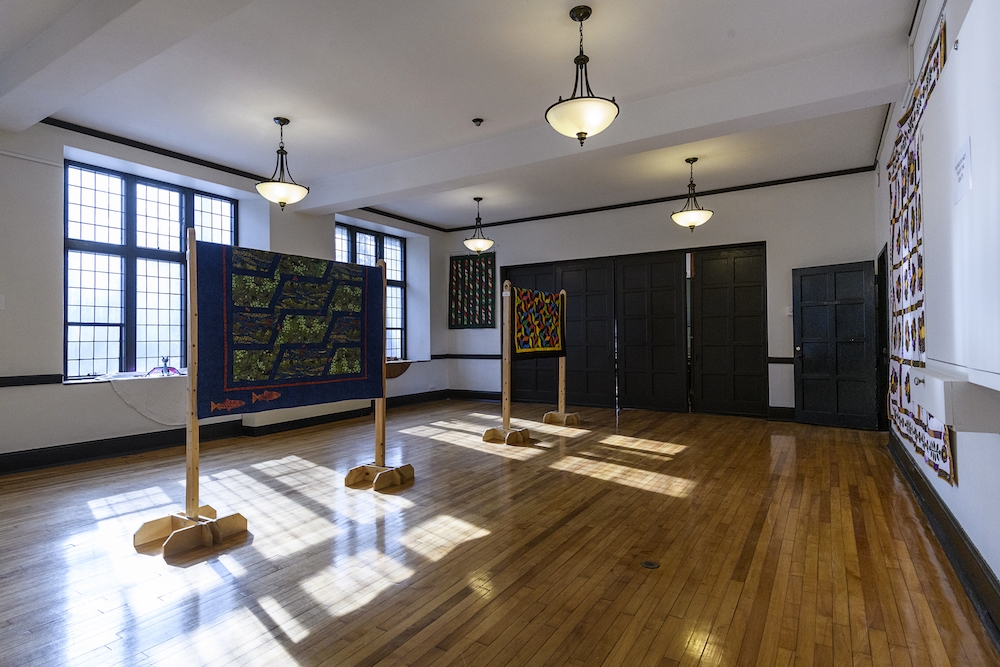 A sun-filled room housing several custom knitted quilts on display for an art exhibit