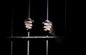 A pair of hands in the dark holding onto the bars of a prison cell.