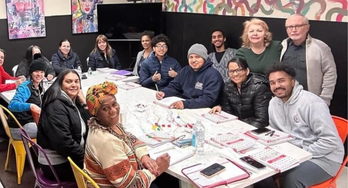 A large group of adults sitting at a long rectangular table working on paper work in binders and notebooks, turning their heads to smile and pose for a photo