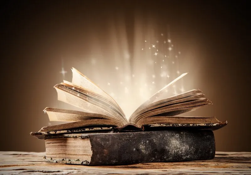 Two old and tattered books laying on a table, the one on top of the other one is open with sparkling light coming from it.