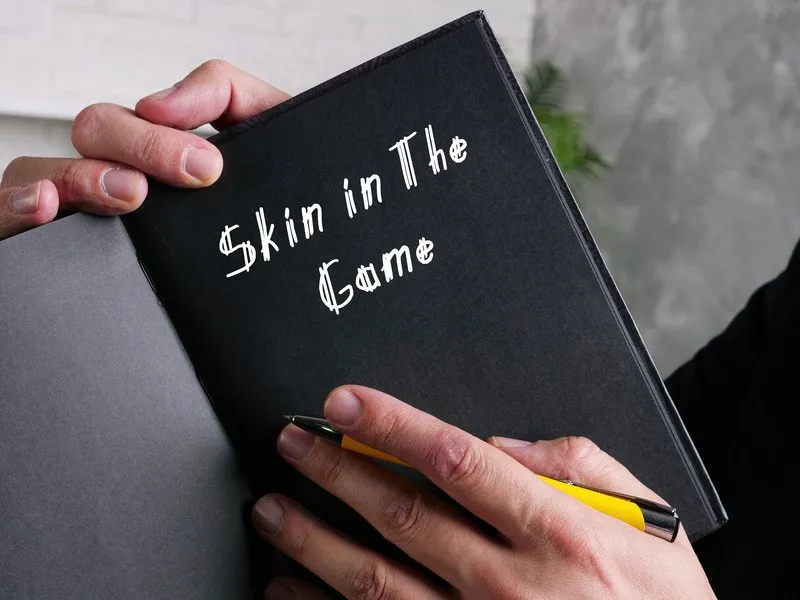 A person holds up a black book with "Skin in the Game" written inside it, while holding a pencil.