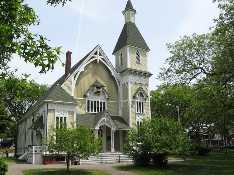 the exterior view of a yellow church with trees and bushes surrounding and a cloudy blue sky in the background