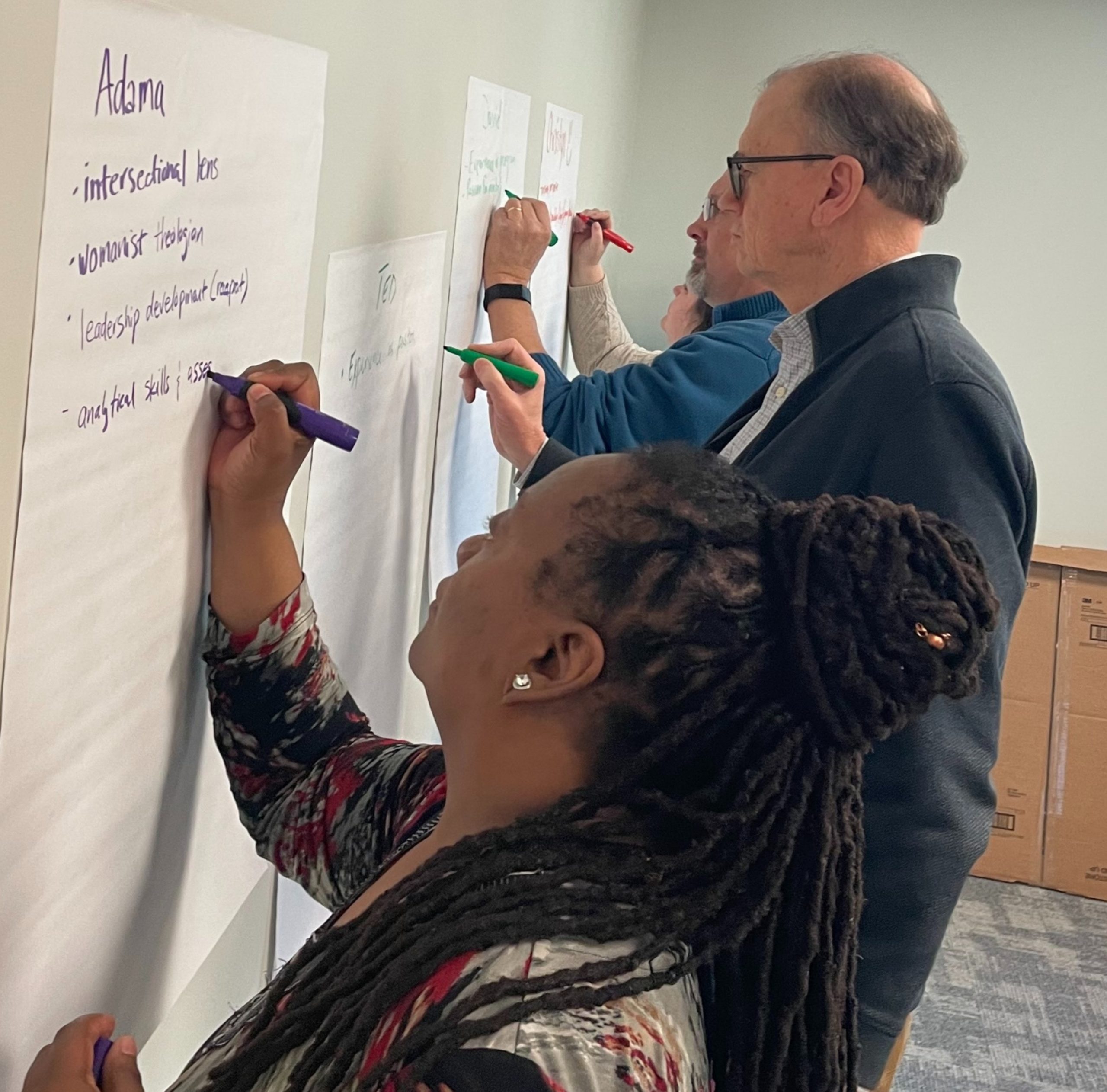 A woman and two men writing with markers on large poster paper taped to the wall