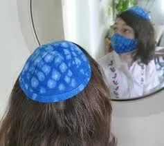 A young man wearing a yarmulke and a face mask looking into a mirror.